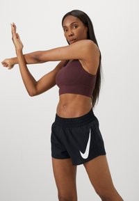 Soutien-gorge de sport maroon et shorts noirs avec le logo blanc de Nike. Le soutien-gorge a une texture lisse ; les shorts présentent une taille élastique et une coupe ample.