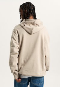 Felpa beige con cappuccio e vestibilità rilassata, caratterizzata da maniche lunghe e una texture morbida. Indossata sopra jeans chiari.