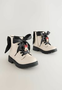 Helle beige Knöchelstiefel mit schwarzen Satinschnürsenkeln, jeweils mit einem runden Minnie-Maus-Anhänger an jedem Stiefel und einer strukturierten schwarzen Sohle.