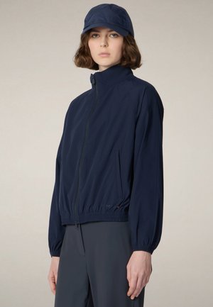 Frau trägt eine marineblaue Jacke mit Reißverschluss, passende Kappe und dunkle Hose, steht vor einem schlichten hellen Hintergrund.