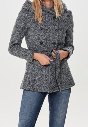 Kobieta w szarym, teksturowanym, dwurzędowym płaszczu z guzikami i wiązaniem w talii, w połączeniu z niebieskimi dżinsami, stojąca na jednolitym tle.