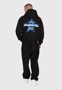 Man in een zwarte hoodie met een blauwe ster en witte tekst "Dropsize Clo" op de achterkant, zwarte broek en witte sneakers, met zijn rug naar de camera gekeerd.