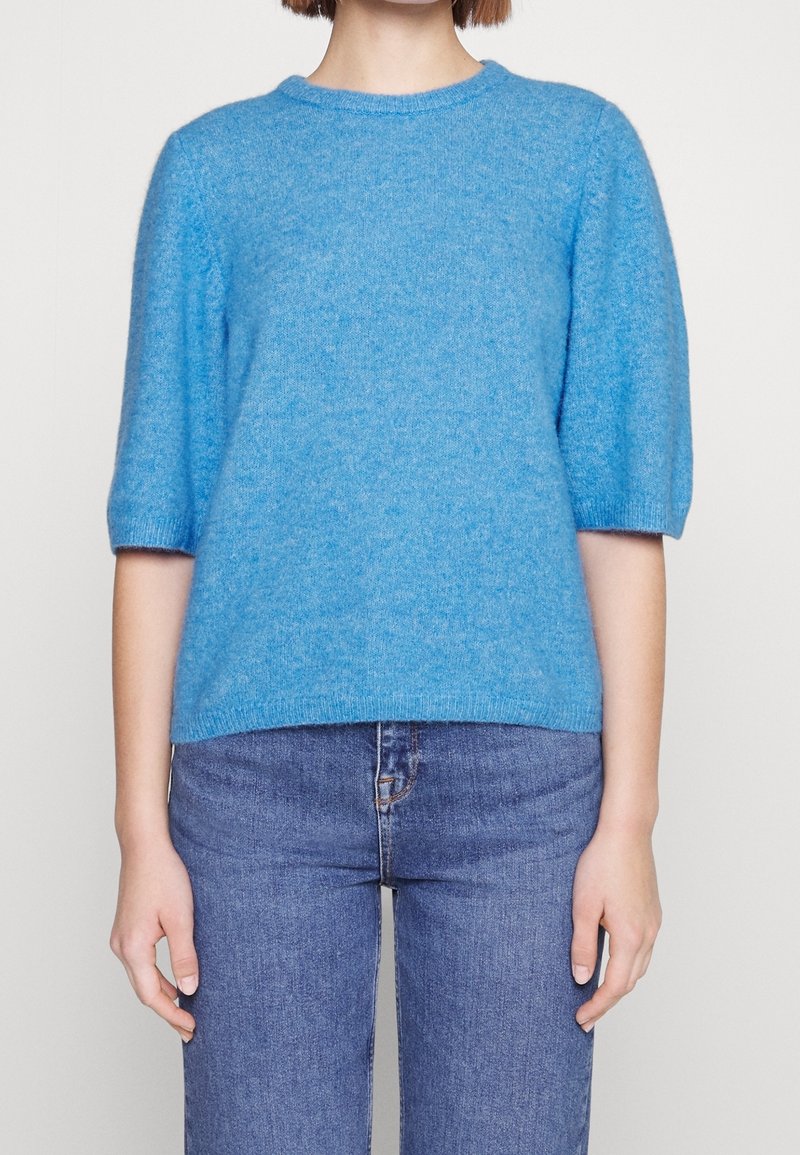 Personne portant un pull bleu doux à manches courtes associé à un jean bleu moyen taille haute, montré du cou aux hanches.