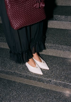 Person trägt schwarze Rüschenhosen und weiße spitze Schuhe mit Perlendetail, hält eine große strukturierte dunkelrote Handtasche und steht auf Steinstufen.