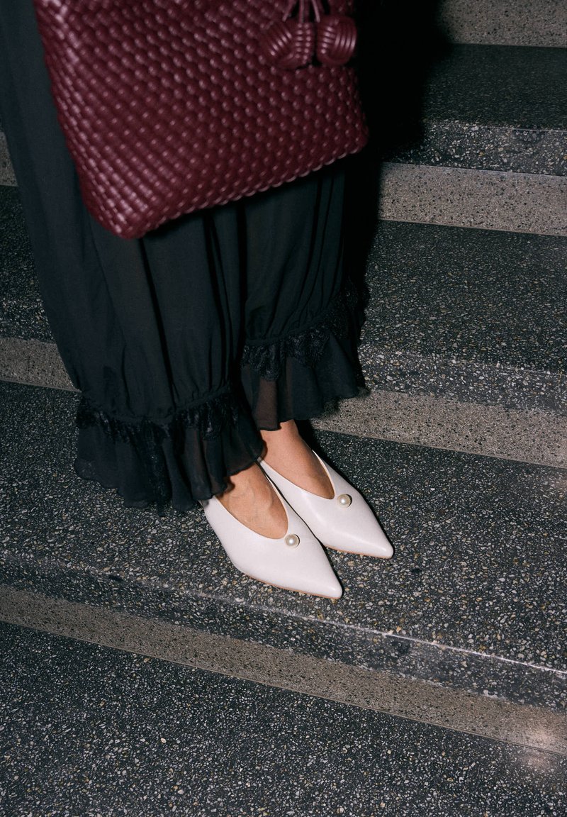 Personne portant un pantalon noir à volants et des chaussures blanches pointues avec détail en perle, tenant un grand sac à main texturé rouge foncé, debout sur des marches en pierre.
