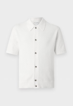 Jonge man met krullend haar die een wit kortermouwig overhemd met knopen draagt, olijfgroene broek en zwarte loafers, staand met handen in de zakken.