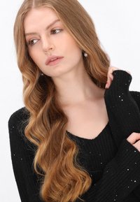Fekete bordázott pulóver V-nyakkal és hosszú ujjú. Apró flitterek díszítik az anyagot. Hosszú, hullámos barna haj keretezi az arcot.