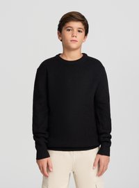 Czarny sweter dziany z prążkowaną fakturą, z okrągłym dekoltem i długimi rękawami, noszony na jasnobeżowych spodniach.