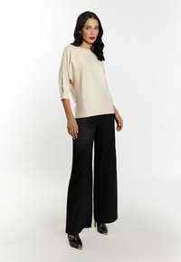 Beige gestricktes Oberteil mit seitlichen Schlitzen und drei Viertel Ärmeln, kombiniert mit schwarzen weit geschnittenen Hosen und schwarzen Pumps mit goldenen Akzenten.