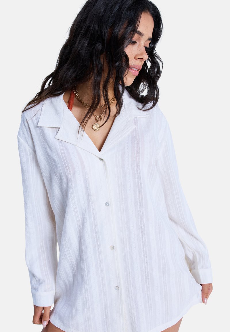 Biała, teksturowana koszula zapinana na guziki z kołnierzykiem, długimi rękawami i subtelnym paskowym wzorem. Posiada przezroczyste guziki i luźny krój.