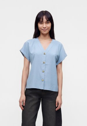 Femme aux longs cheveux foncés porte un haut bleu clair à manches courtes boutonné et un pantalon noir à jambes larges, debout devant un fond uni.