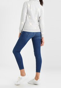 Sudadera gris con patrón de pájaros en crema, combinada con jeans ajustados azules y zapatillas blancas. Presenta un cuello alto y puños acanalados.