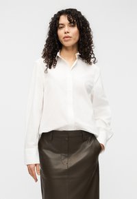 Λευκό πουκάμισο με μακριά μανίκια και κλασικό γιακά. Συνδυάζεται με εφαρμοστή φούστα από σκουρόχρωμο καφέ δέρμα. Λεπτή υφή.