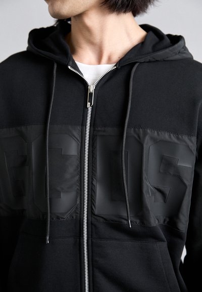 Schwarze Kapuzenjacke mit Reißverschluss, Fronttaschen und einem markanten matten Finish-Paneel mit geprägten Buchstaben. Weicher Stoff und verstellbare Kordel.