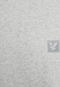 Hellgrauer Sweatshirt-Stoff mit weicher Textur. Enthält ein kleines, geprägtes Adler-Logo in der oberen linken Ecke.