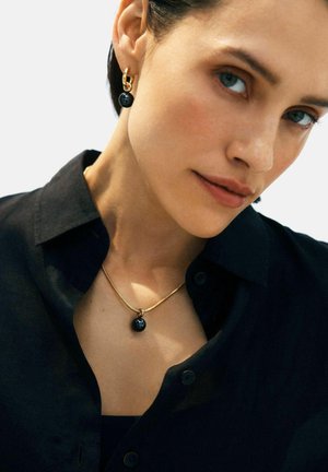 Žena koja nosi zlatne obruče i zlatnu ogrlicu s crnim kuglastim privjescima, odjevena u crnu košulju s gumbima.