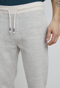 Pantalones de chándal de color gris claro con un tejido texturizado, cordón blanco y cintura elástica. La vista lateral muestra el ajuste y la colocación de las manos.