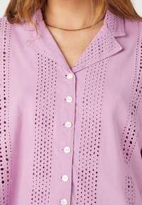 Lawendowa bluzka z haftem ażurowym, białymi guzikami z przodu, kołnierzykiem i cienkim łańcuszkiem złotym na szyi osoby.