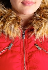 Červená vatovaná bunda s umělým kožešinovým límcem. Má stříbrné zipy a texturovanou látku. Detailní záběr na límec a zipy.