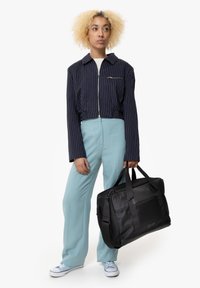 Musta kassi tekstuuriga detailidega ja kahe käepidemega, mida hoiab inimene, kellel on seljas tumesinise triibuline jakk ja heledad sinised püksid.