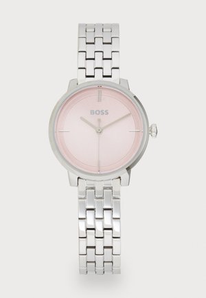 Montre bracelet en argent avec un cadran rose, dotée d'un design minimaliste, de marqueurs d'heure en argent et d'un bracelet à maillons en métal. Logo de la marque affiché.