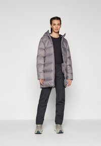 Manteau rembourré gris avec capuche, présentant des sections matelassées. Associé à un pantalon foncé et des chaussures lacées gris clair. Texture lisse.
