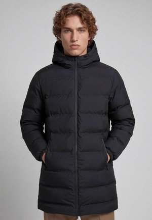 Jovem com cabelo encaracolado vestindo um casaco acolchoado preto comprido com capuz, mãos nos bolsos, em pé contra um fundo cinza liso.