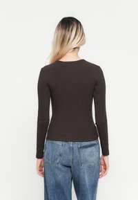 Kvinne med langt blondt hår iført en tettsittende mørk brun langermet skjorte og løse blå denimjeans, vendt bort fra kameraet mot en ensfarget hvit bakgrunn.