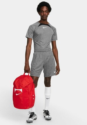Maglietta atletica grigia e pantaloni corti con dettagli neri, abbinati a un grande zaino rosso Nike. Calzini bianchi e scarpe da calcio nere completano l'outfit.