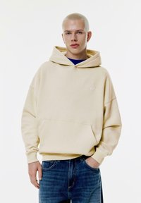 Beige hoodie med en framficka och ribbade ärmmuddar, med en liten logotyp. Bärs över en blå skjorta och matchas med blå jeans.