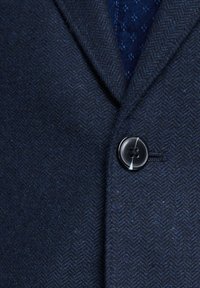 Blazer a spina di pesce blu navy con un aspetto texture; presenta un bottone rotondo nero con quattro fori e un accento bianco a contrasto.