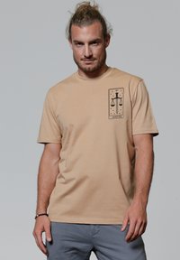 Beige katoenen t-shirt met korte mouwen, voorzien van een zwart grafisch ontwerp van schubben en een zwaard op de linkerkant van de borst. Soepele textuur.