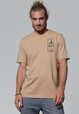 Beige cotton t-shirt with short sleeves featuring a black graphic design of scales and a sword on the left chest. Smooth texture.