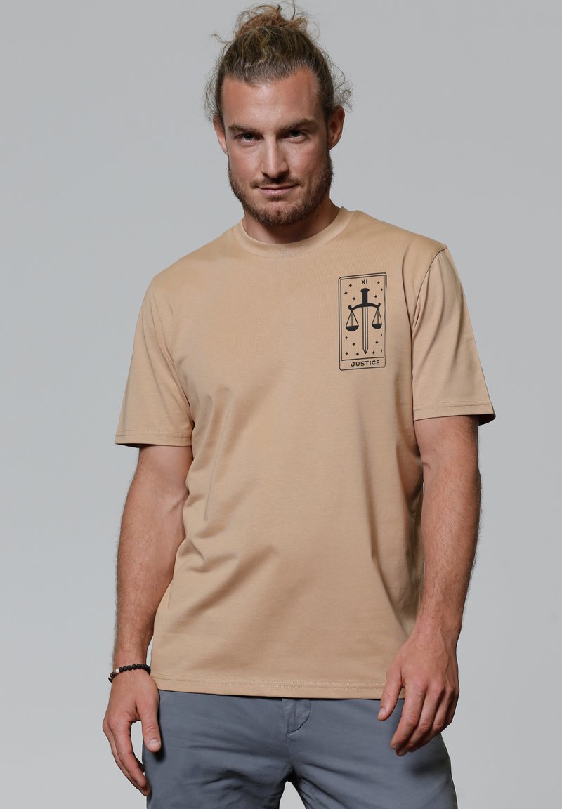 Beige katoenen t-shirt met korte mouwen, voorzien van een zwart grafisch ontwerp van schubben en een zwaard op de linkerkant van de borst. Soepele textuur.