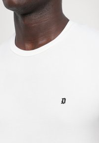Personne à la peau foncée portant un t-shirt blanc uni à col rond avec une petite lettre D noire sur le côté gauche de la poitrine.
