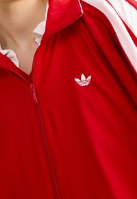 Röd zip-up träningsjacka med vita ränder, med en vit broderad logotyp på vänster bröst. Slät textur, avslappnad design.