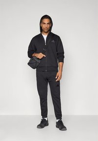 Roupa desportiva preta composta por um hoodie com fecho e calças de treino, com o logótipo da Adidas. Acompanhada por uma pequena bolsa transversal e sapatilhas.