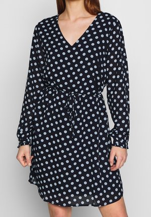 Donna che indossa un vestito blu navy a maniche lunghe con pois bianchi e cintura in vita, scollatura a V.