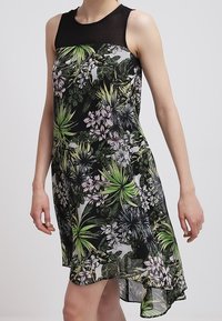 Květinové šaty s černým korzetem, průhlednými ramenními panely a splývavou dolní částí. Zelený a fialový botanický potisk na světlém pozadí.