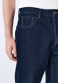 Granatowe dżinsy o gładkiej fakturze, widocznym przeszyciu, standardowym pasku i bocznymi kieszeniami. Bez przetarć ani ozdób.