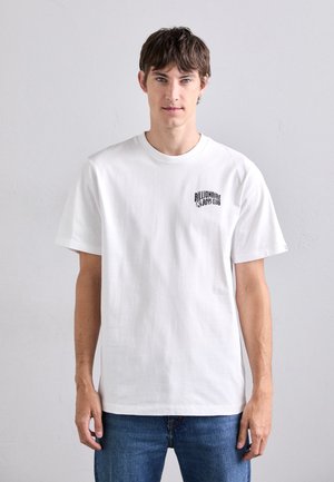 White cotton t-shirt with a crew neck and short sleeves, featuring a black printed logo on the left chest. Worn with blue jeans.