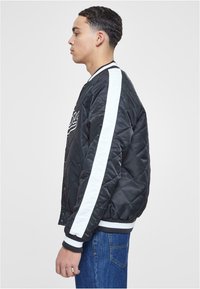 Black quilted bomber jacket with a white stripe down the sleeve, ribbed cuffs, and a front zip. Features a logo detail on the chest.