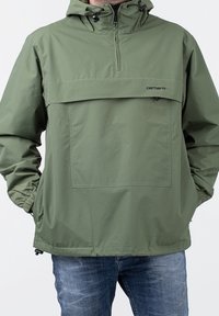 Letvægts grøn pullover-jakke med halvlynlås foran, justerbar hætte, stor frontlomme og elastiske manchetter. Fremstillet af vandafvisende materiale.