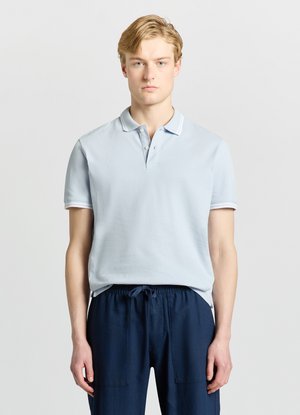 Junger Mann mit blondem Haar, der ein hellblaues Poloshirt und marineblaue Hose mit Kordelzug trägt, steht vor einem einfachen hellen Hintergrund.