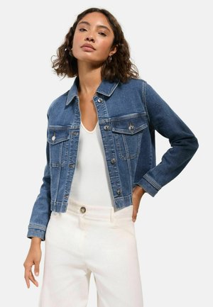 Frau trägt blaue Jeansjacke über weißem Oberteil und weißen Hosen, posiert mit einer Hand in der Hüfte vor einem einfarbigen Hintergrund.