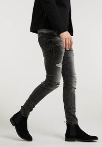 Persona caminando, vistiendo botas Chelsea de ante negras, jeans ajustados de color gris oscuro desgastados y una chaqueta negra con botones.