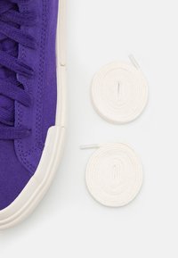 Purple canvas sneaker with textured upper, white rubber sole, and two coiled white laces positioned beside it.