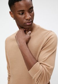 Suéter de punto beige con un patrón texturizado, cuello y puños acanalados. Mangas largas y silueta ajustada, destacando la suavidad de la tela.