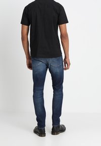 Czarna koszulka z krótkim rękawem, niebieskie dżinsy o zwężanym kroju i czarne buty. Widoczne szwy i subtelne detale tylnych kieszeni.