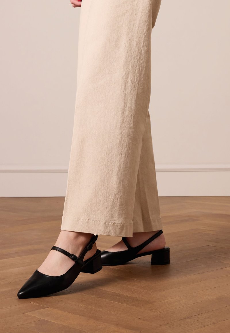 Zwarte leren slingback schoenen met een spitse neus, kleine blokhak en enkelriem. De achtergrond toont lichtgekleurde stoffen broek.